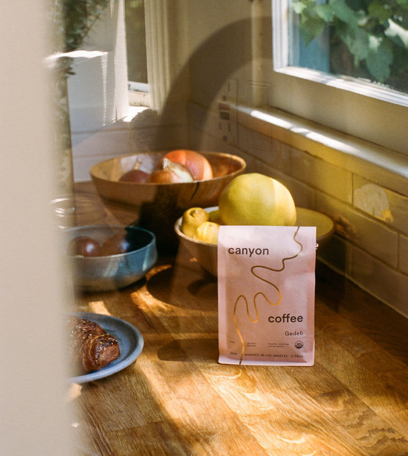 A pink bag of Canyon Coffee on a wood counter with a bowl of fruit
